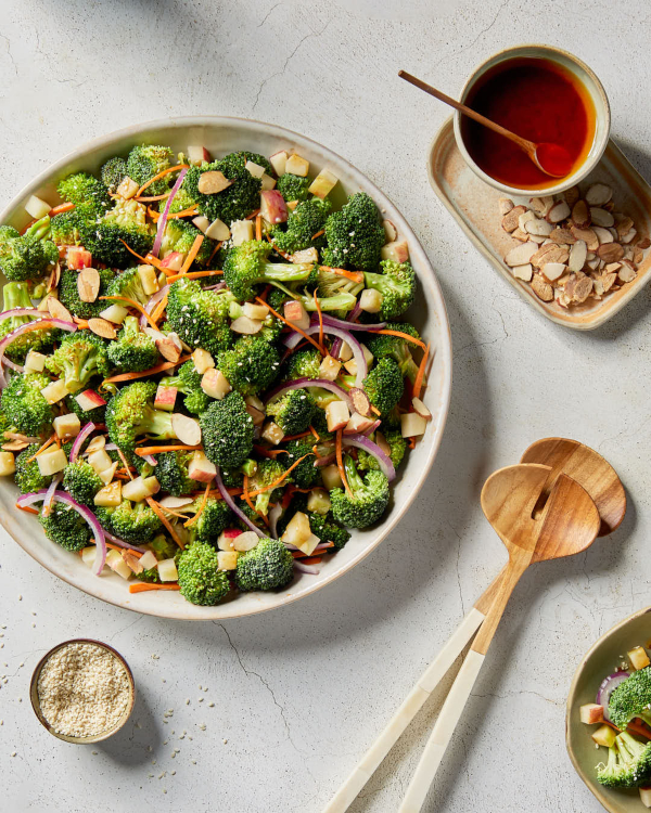 A serving bowl of broccoli salad with gochujang dressing on a kitchen counter shown with a pitcher of dressing, and wooden serving spoons.