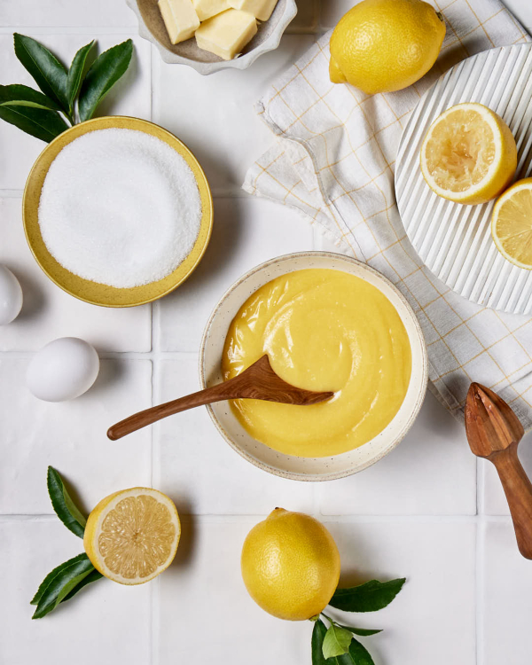 A bowl of lemon curd shown on a tiled counter with whole and halved lemons, a lemon juicer, and a bowl of granulated sugar.