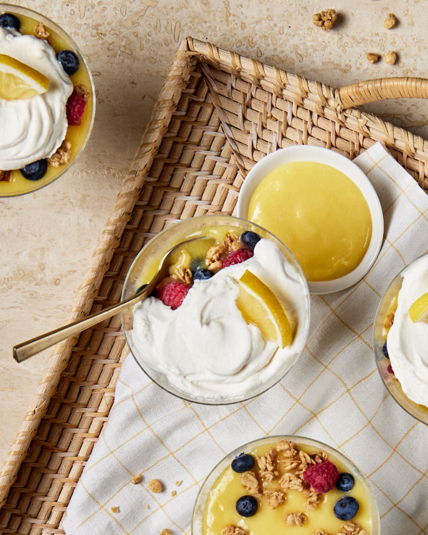 A glass dish of lemon parfait shown on a wicker tray with a bowl of lemon curd and three other parfaits. 