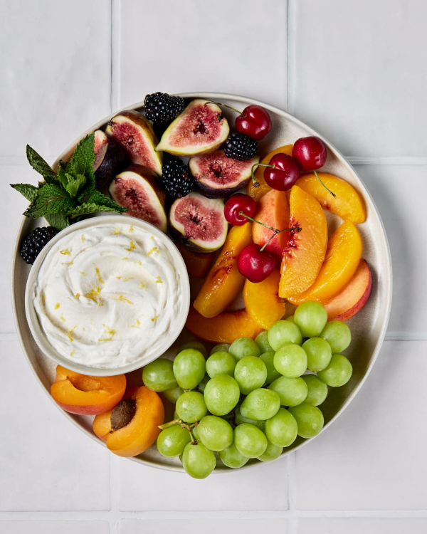 A fruit platter on a tiled counter with a bowl of lemon whipped cream, figs, blackberries, peaches, cherries, and grapes.