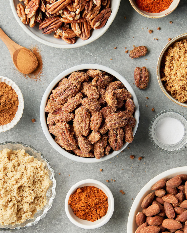A white bowl overflowing with candied pecans and almonds, shown on a kitchen counter with a wooden spoon and bowls of plain almonds and pecans, brown sugar, cinnamon, and paprika