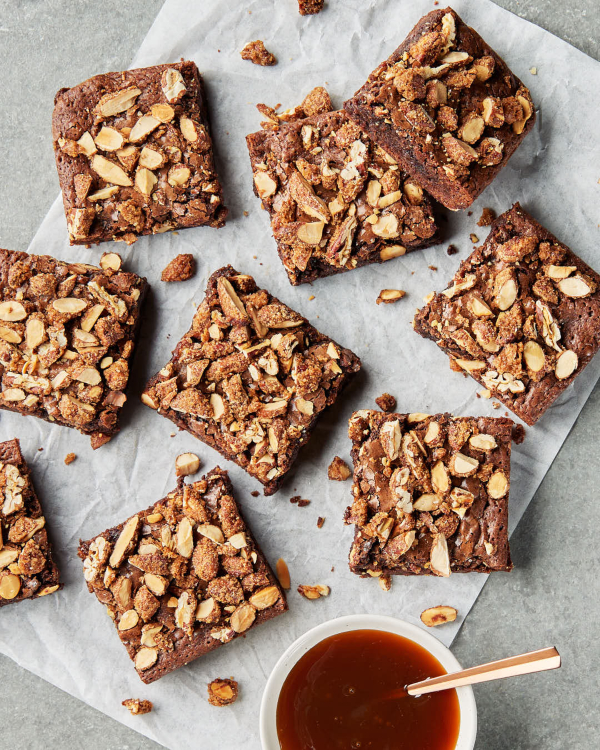 Cut chocolate brownies topped with candied nuts and caramel sauce, shown on a piece of waxed paper on a kitchen counter with a bowl of caramel sauce.
