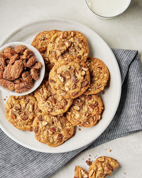 A plate of homemade cookies with chopped candied nuts, shown with a bowl of candied nuts and a glass of milk.