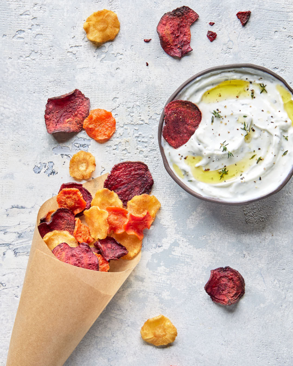 A parchment paper cone full of mixed air fryer root vegetable chips with several chips scattered around, shown with a bowl of creamy dip.
