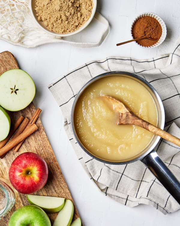 A small saucepan of applesauce with a wooden spoon dipped in, shown on a tea towel with whole and sliced apples, cinnamon sticks, a bowl of cinnamon, and a bowl of Golden Yellow Sugar.