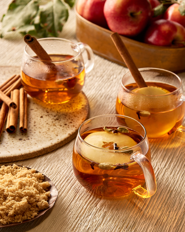 Three glass mugs of hot apple cider garnished with apple slices, cloves, and cinnamon sticks, shown on a table with a pitcher of cider, a bowl of apples, and a bowl of golden yellow sugar 