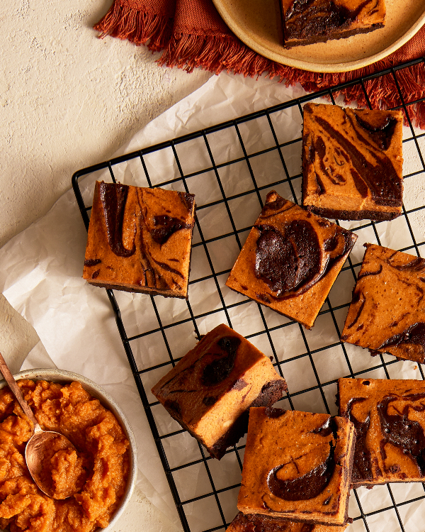 Seven pumpkin cheesecake brownies on a wooden cutting board lined with parchment, shown with a baking pan with brownies and a bowl of pumpkin puree.
