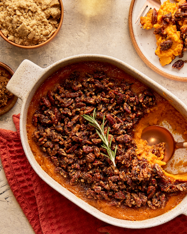 A partially served sweet potato casserole garnished with a rosemary sprig in a baking dish, shown with a serving spoon, a plate of casserole, and bowls of golden yellow and demerara sugar.