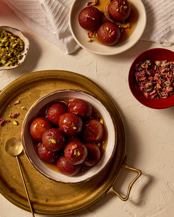 Balls of fried dough (Gulab Jamuns) covered in syrup on a golden plate with a golden spoon and plates of rose petals and pistachio 