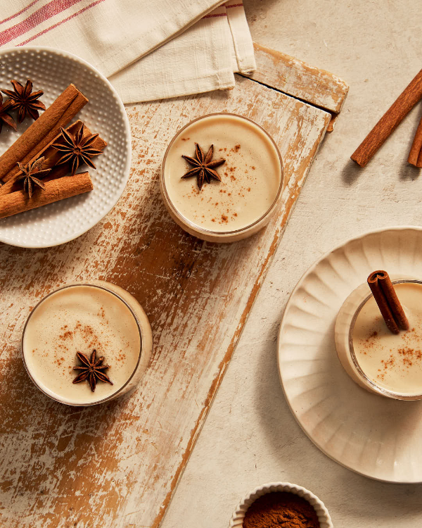 Two tumbler glasses of eggless eggnog garnished with anise, cinnamon, on a rustic wooden cutting board with a third glass on a saucer, shown with cinnamon and anise in a dish.