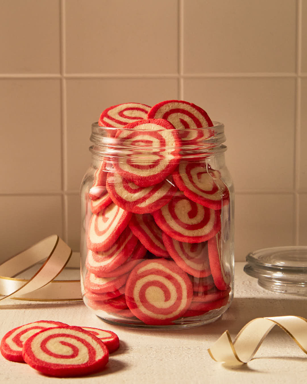 A tall glass jar full of red and white peppermint pinwheel cookies on a kitchen counter in front of a cream tile backsplash, shown with the glass lid, white and gold ribbon, and three cookies on the counter.