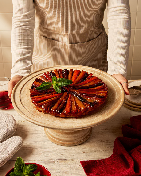 A woman standing behind a A carrot and parsnip tarte tatin on a cake stand, shown with fresh mint leaves, oven mitts, a dark beverage, and serving plates.
