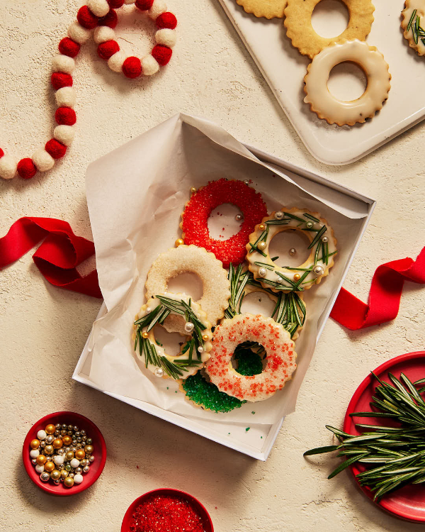 Glazed sugar cookies with rosemary shown decorated in various winter holiday themes, in a gift box with a red ribbon for wrapping, shown with more cookies on a tray, fresh rosemary, and holiday decorations.