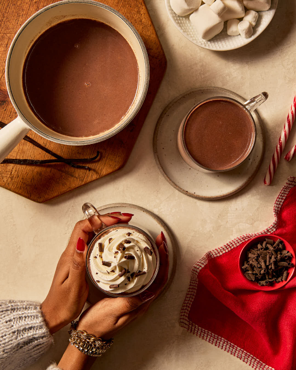 Two feminine hands holding a glass mug of vanilla hot chocolate topped with whipped cream and garnished with shaved chocolate, resting on saucers and shown with marshmallows, shaved chocolate, candy canes, and another mug of hot chocolate.