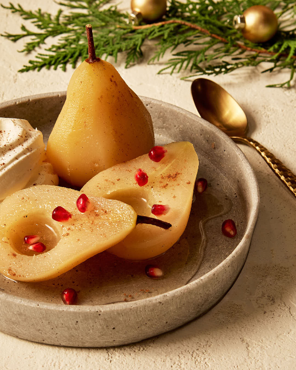 A white wine poached pear cut in half in a shallow dish, served with a dollop of whipped cream and sprinkled with cinnamon and pomegranate arils, and shown with a gold spoon, a red dish of pomegranate arils, and a small pot of halved pears soaking with seasonings.