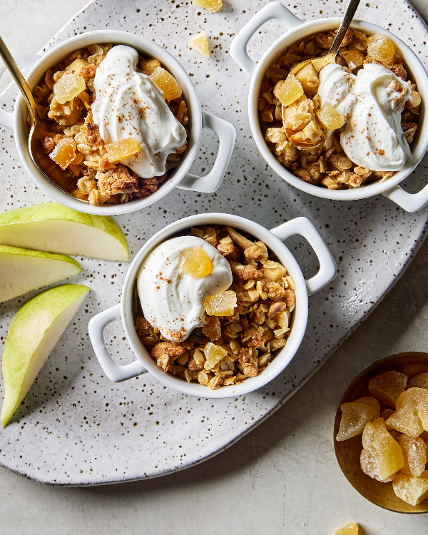 Overhead view of three white single-serving bowls of pear ginger crumble with whipped cream, two with gold spoons, shown with pear slices and a small dish of ginger pieces.