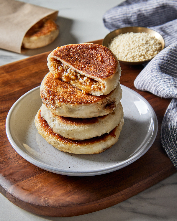 Five Korean pancakes or hotteok, four stacked on a plate on a wood tray and one wrapped in paper, with one cut open with sesame syrup leaking out, shown with a bowl of sesame seeds.