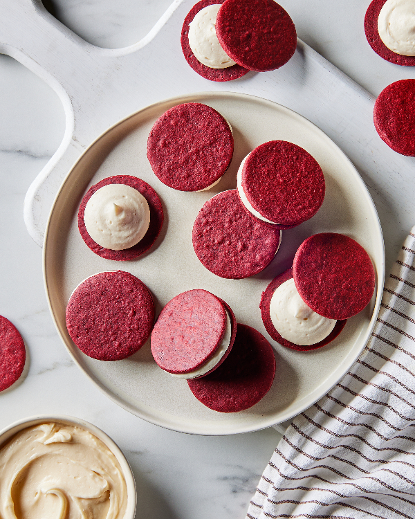 A plate of red velvet sandwich cookies with cream filling on a marble countertop, next to a striped kitchen towel and a bowl of extra cream filling