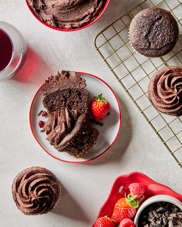Red wine chocolate cupcakes, some plain, some topped with ganache, with two on a wire cooling rack, one on a counter and one cut in half on a plate, shown with a bowl of ganache, shaved chocolate, and strawberries.