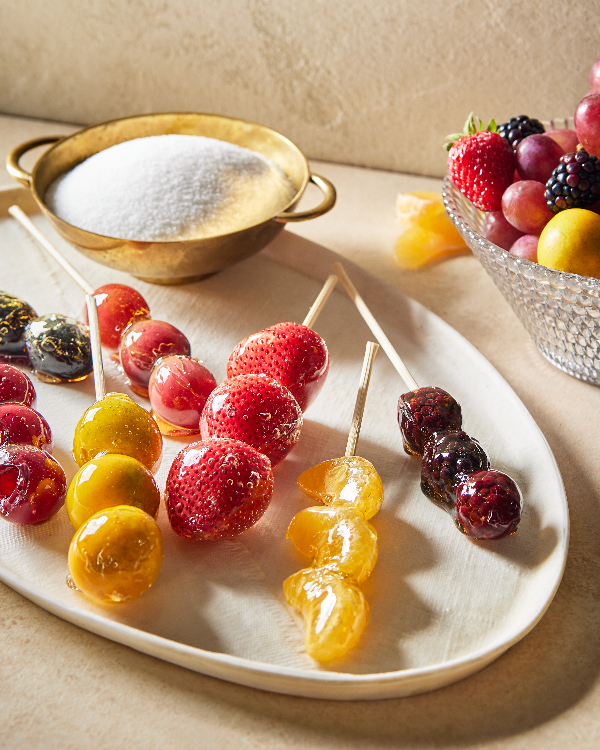  A platter of skewered fruit and berries coated in clear sugar syrup, or tanghulu, including mandarin segments, cherries, grapes, gooseberries, strawberries, kumquats, and blackberries, shown with a bowl of fruit and a bowl of granulated sugar.