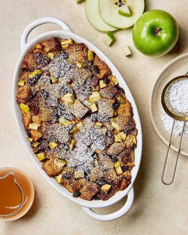 An oval baking dish full of apple bread pudding shown dusted with icing sugar accompanied by a glass pitcher of butterscotch sauce and sliced and whole green apples.