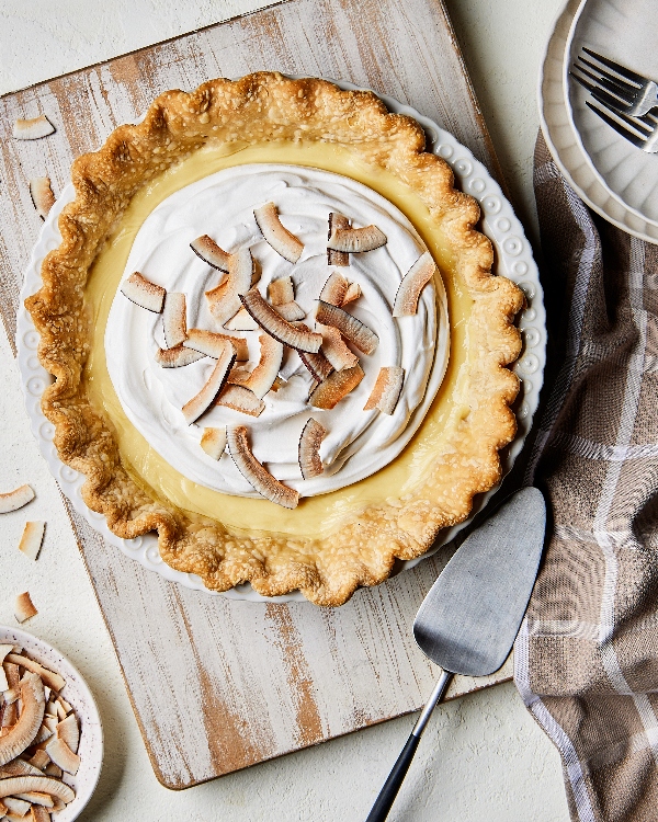 Freshly baked coconut cream pie with golden-brown flaky crust and creamy topping, garnished with toasted coconut flakes, served on a classic white pie dish, accompanied by a pie server on a rustic wooden tabletop.