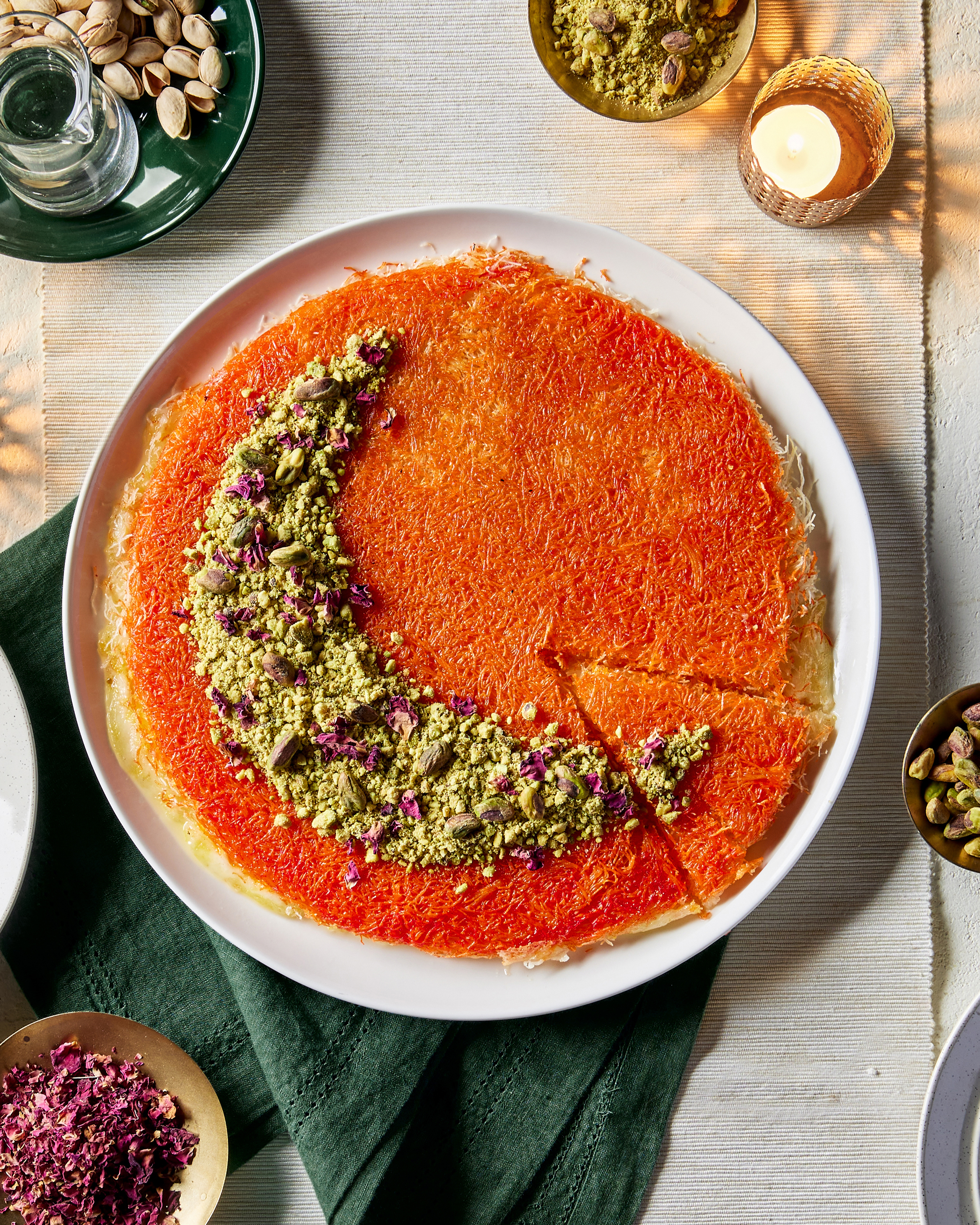 Knafeh on a plate with one slice cut but not removed, garnished with pistachios and flower petals arranged in a crescent moon shape, shown with dishes of pistachios in the shell and flower petals and a lit candle. 