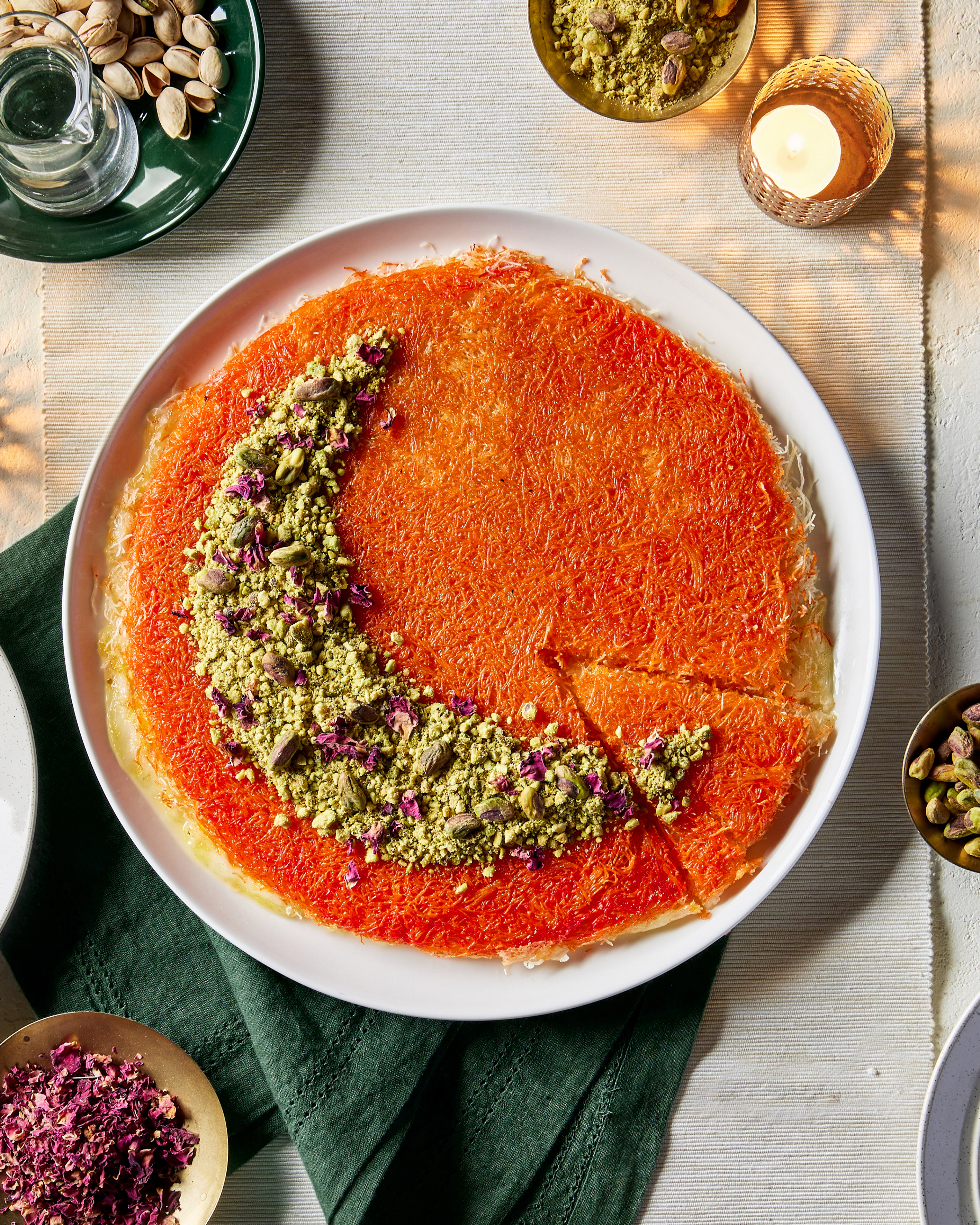 Knafeh on a plate with one slice cut but not removed, garnished with pistachios and flower petals arranged in a crescent moon shape, shown with dishes of pistachios in the shell and flower petals and a lit candle. 