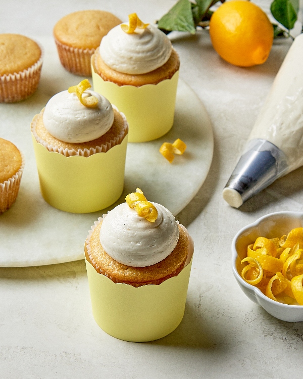 Three lemon cupcakes with creamy frosting and lemon peel garnish and three more without toppings displayed on a marble countertop with a filled piping bag and fresh lemons in the background.