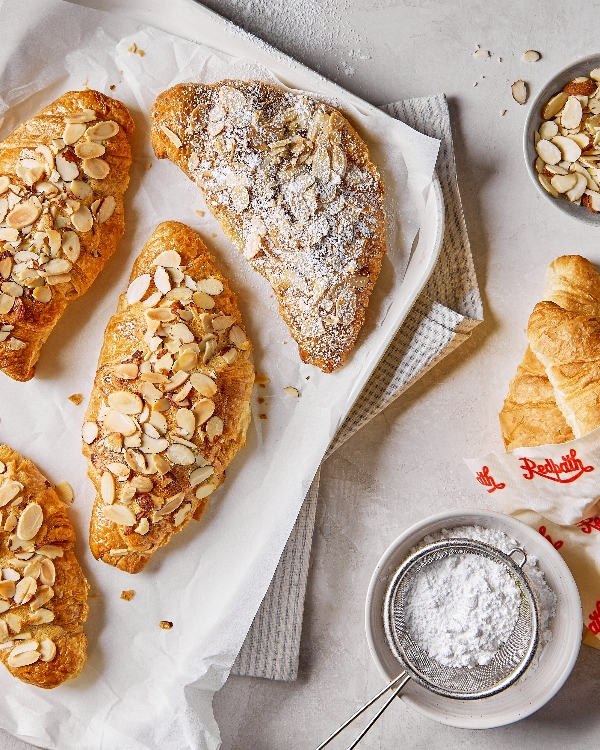 Overhead view of three air fryer almond croissants with a generous topping of sliced almonds and powdered sugar on parchment paper, with an additional plain croissant and a sifter full of powdered sugar on a marble surface.