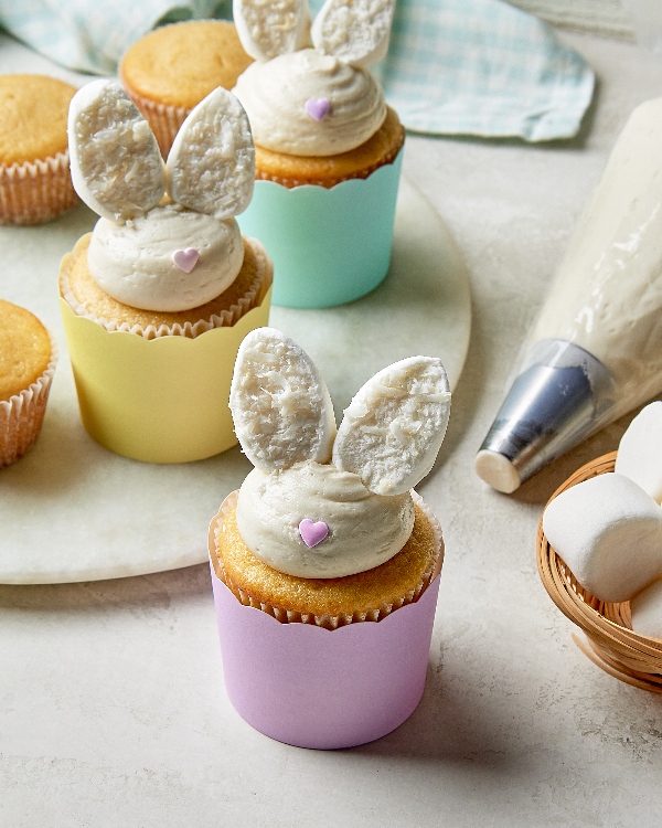 A close-up image of a decorated cupcake designed to look like a bunny. The cupcake has two bunny ears made of cut and textured marshmallows positioned in a swirl of cream-colored frosting on top. It's placed in a purple cupcake wrapper and has a small pink heart-shaped sprinkle in the center of the frosting. In the background, there are more cupcakes, a pastry bag, and marshmallows, all on a marble countertop.