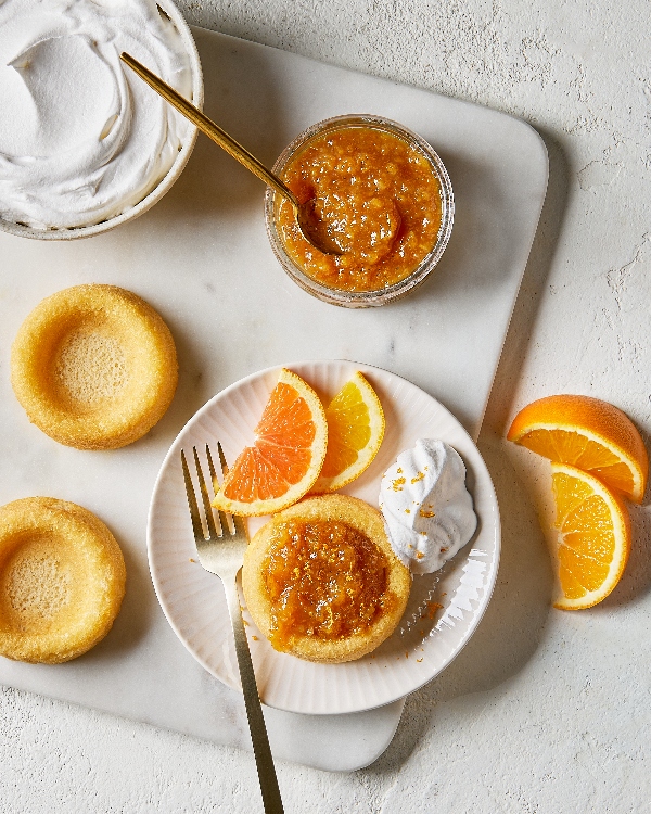 Top-down view of a dessert setting featuring a sponge cake with orange marmalade topping and a side of whipped cream, fresh orange slices on the plate, extra sponge cakes, and a bowl of marmalade with a gold spoon, on a marble countertop.