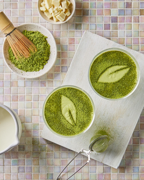 Overhead view of two bowls of white chocolate matcha mousse topped with a fine dusting of matcha powder, alongside ingredients like white chocolate chunks and a jug of cream, with a bamboo whisk and a sieve on a white wooden board, all set upon a multicoloured tiled surface.