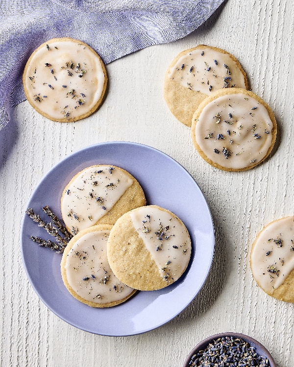 A batch of glazed lemon-lavender shortbread cookies sprinkled with dried lavender, shown with some on a plate and others on a textured surface, with dried lavender stalks, a bowl of dried lavender, and a lavender-coloured cloth.