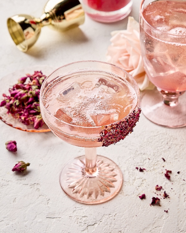 Two unique stemmed glasses of lychee-rose spritzer on ice garnished with dried rose petals on the glasses, shown in a neutral setting with a fresh rose in the background, a plate of dried rose petals, and a gold jigger
