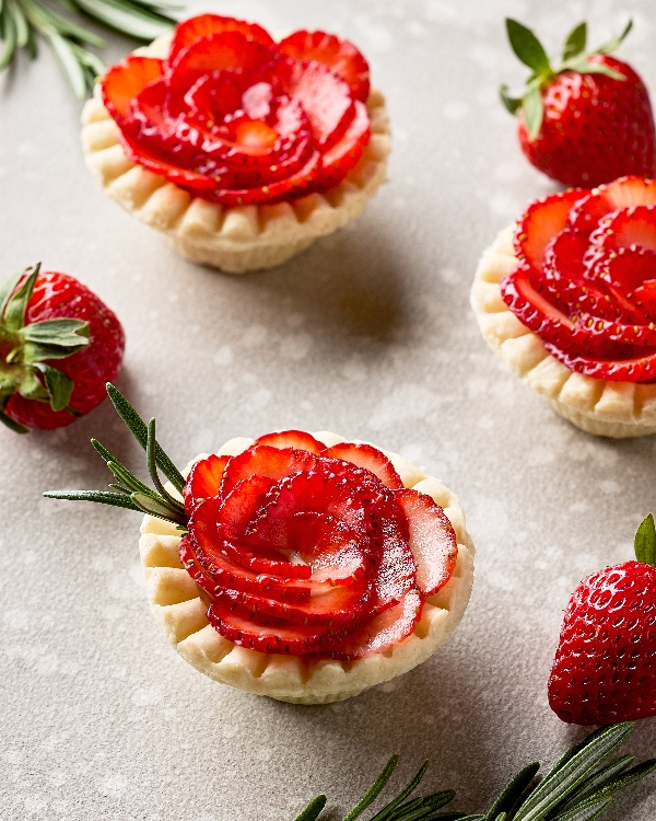 Three strawberry rosemary tartlets on a kitchen counter, shown with three fresh strawberries and sprigs of rosemary