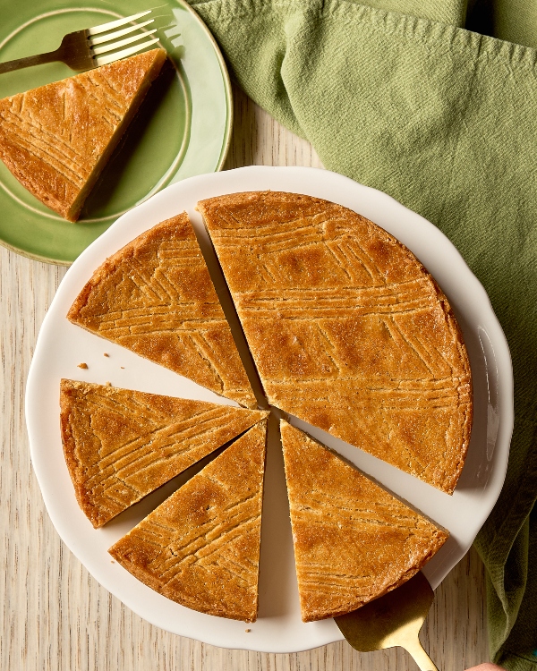 An overhead, close-up shot of a golden-brown, round Brown Sugar Dutch Butter Cake (Boterkoek) cut into six wedges and arranged on a white serving plate. The cake has a dense, buttery texture and features a diamond-patterned score across its surface. A hand using a gold serving spatula is removing one slice from the bottom right. In the upper left, one slice already sits on a small green plate next to a fork, and a crumpled green napkin is next to the plate. The background is a light-colored wooden surface.