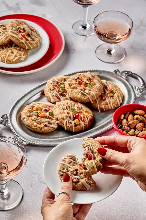 A silver serving tray filled with glazed fruitcake cookies topped with colourful candied fruit. In the foreground, a person breaks a cookie in half over a small white plate. Glasses of rosé and a small bowl of almonds and pistachios sit nearby on a light grey tabletop