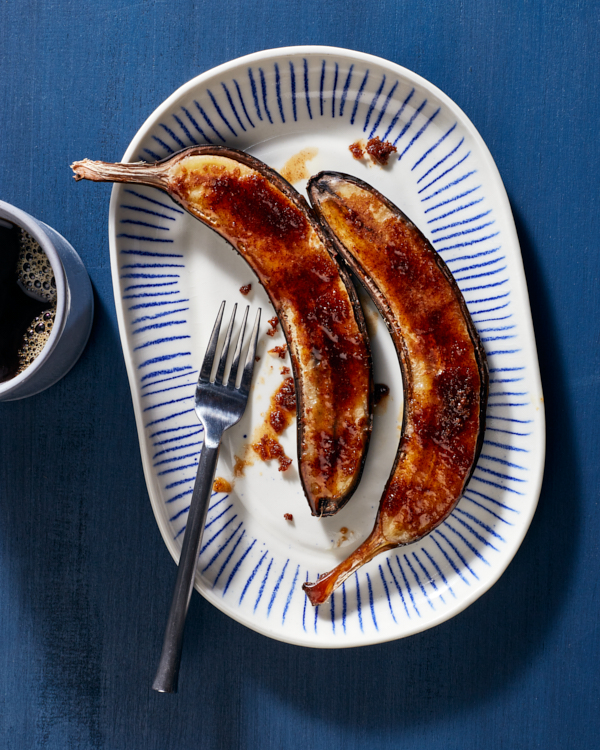  Roasted bananas foster sliced in half lengthwise and topped with brown sugar and cinnamon, served on a blue and white plate with a fork and a mug of coffee.