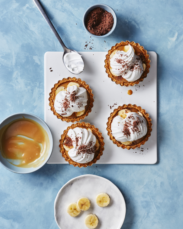 Four single-serving banoffee pies on a white platter shown with a bowl of toffee filling, a bowl of shaved chocolate, and sliced banana on a plate.