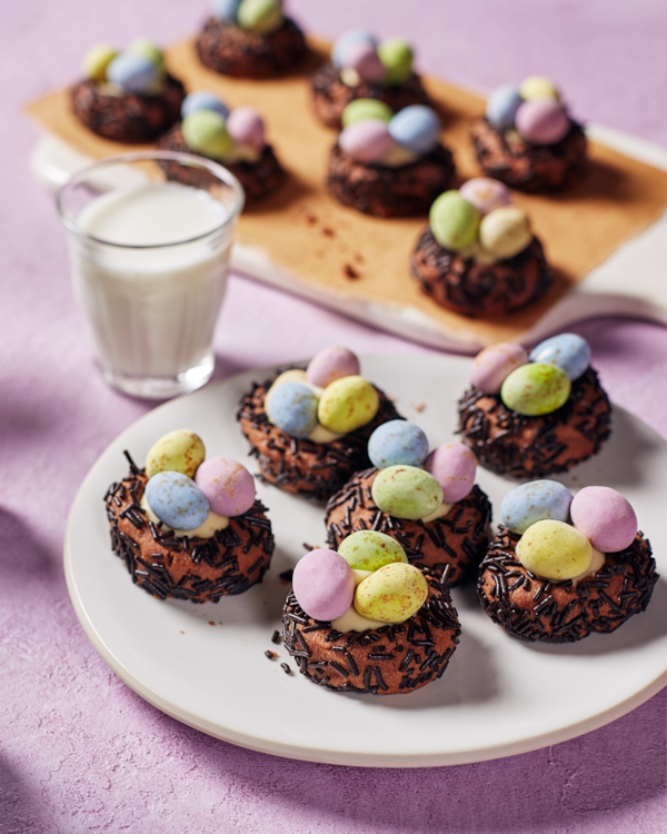 Chocolate bird nest cookies topped with white chocolate and candy-coated chocolate eggs on a plate and a cutting board, shown with a glass of white milk