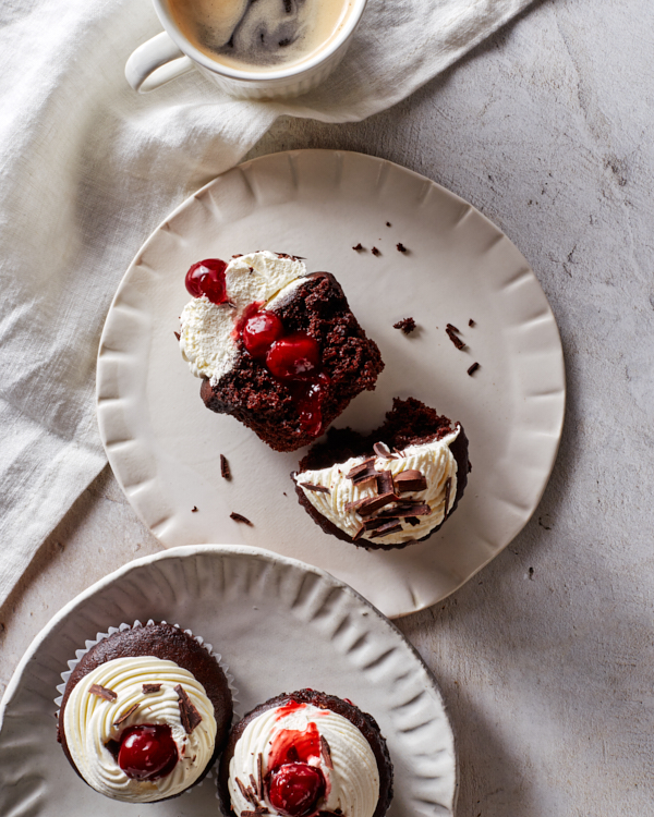 Three Black Forest cupcakes with chantilly icing topped with cherries and shaved chocolate; two on a platter and one cut in half on a plate.