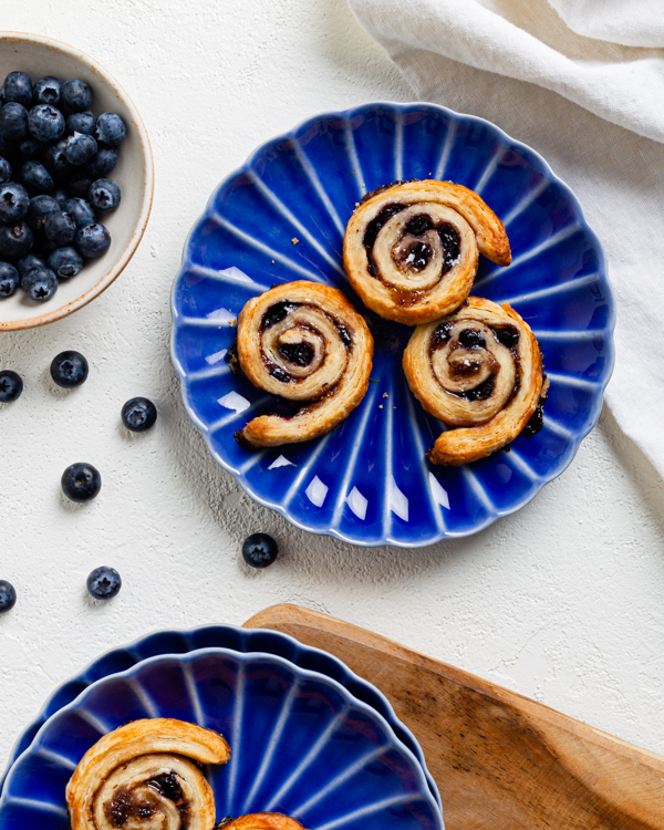 Blue plates with blueberry pie crust pinwheels (pets des soeurs) on a white surface)  with a bowl of blueberries