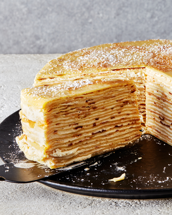 A bourbon crêpe cake dusted with icing sugar on a black platter with one slice being removed on a pie lifter