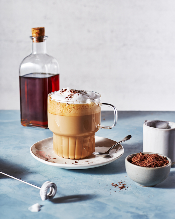 A glass mug full of a frothed latte with whipped topping and shaved chocolate shown with a bottle of brown sugar simple syrup.
