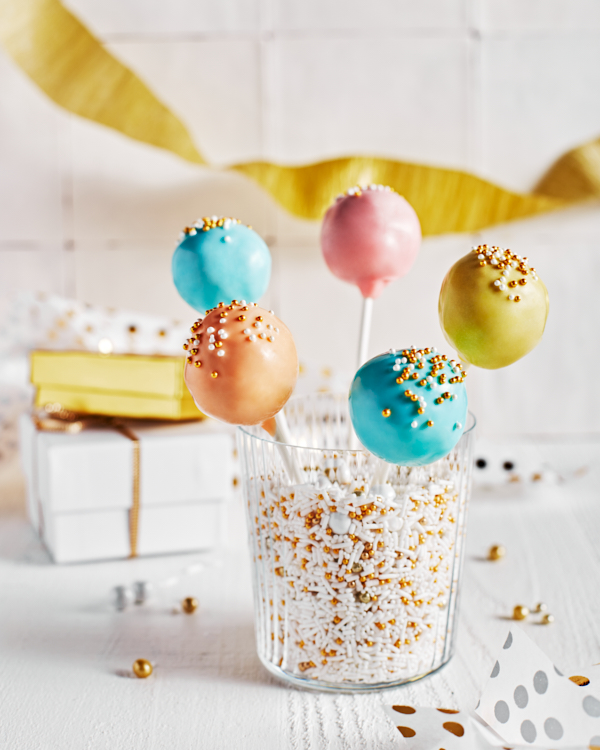 Pastel-coloured cake pops with sprinkles standing in a glass of sprinkles, shown with white and gold party decorations and gifts