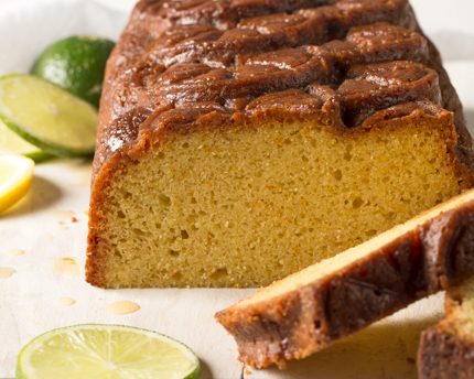 A loaf of pound cake sliced at one end, on a plate with sliced lemons and limes