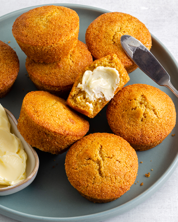  Cornmeal muffins on a plate, one cut in half and buttered, shown with a butter dish and knife