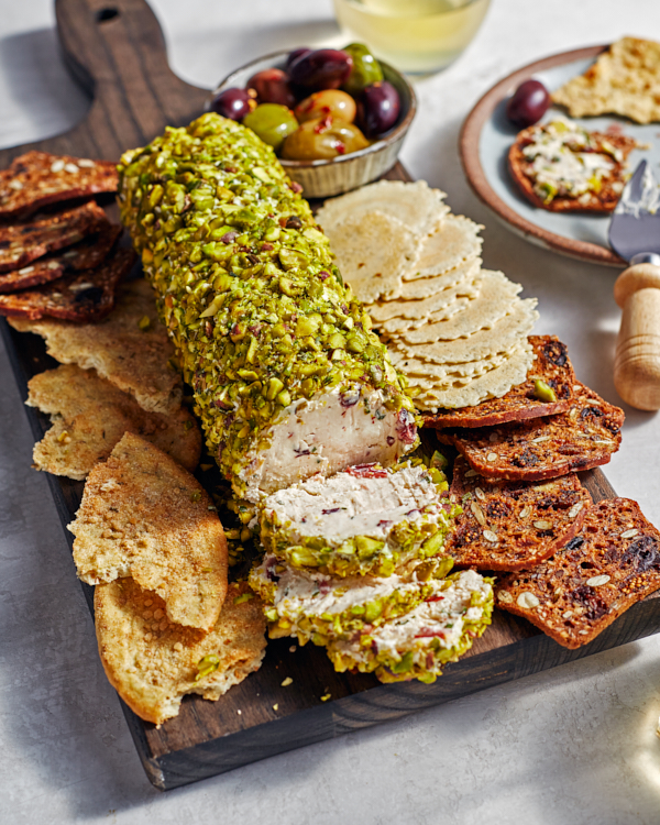 A cream cheese log coated in crushed pistachios on a serving board, surrounded by crisps and crackers and a bowl of olives