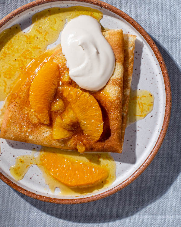 A crepe on a plate topped with beurre Suzette, orange segments, and brown sugar whipped sour cream